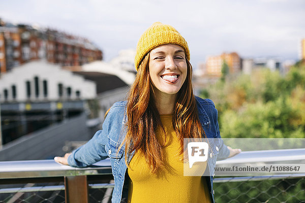 Porträt einer lächelnden jungen Frau mit gelber Mütze und herausstehender Zunge
