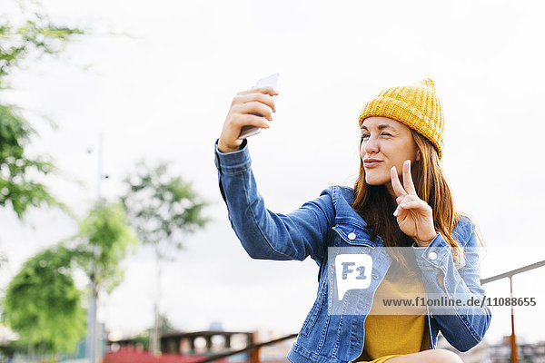 Lächelnde junge Frau mit Siegeszeichen bei der Einnahme von Selfie