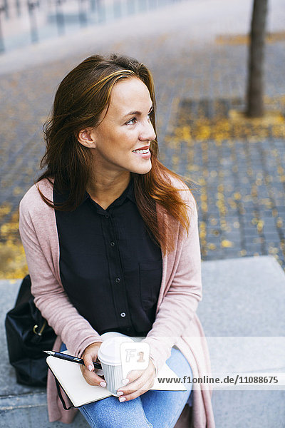 Lächelnde junge Frau sitzt auf Bank mit Notizbuch und Kaffee zum Mitnehmen