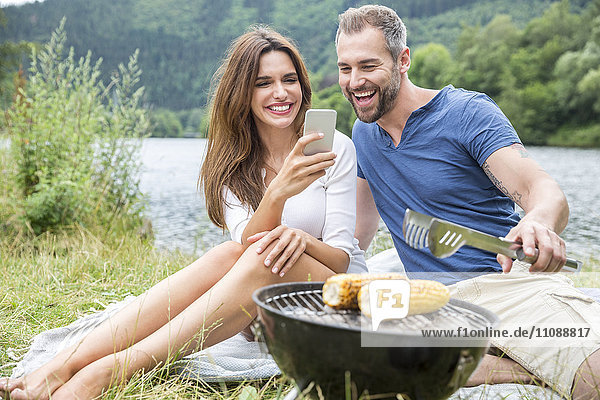 Glückliches Paar mit Handy beim Grillen am Seeufer