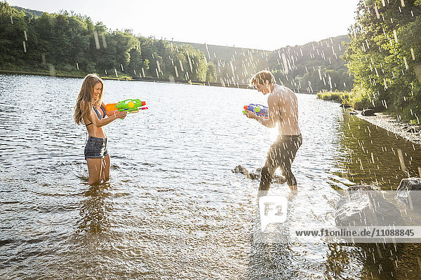 Junges Paar in einem See  das mit Wasserpistolen spritzt.