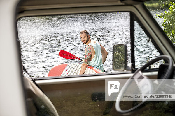 Mann mit Stehpaddelbrett am Seeufer hinter Van