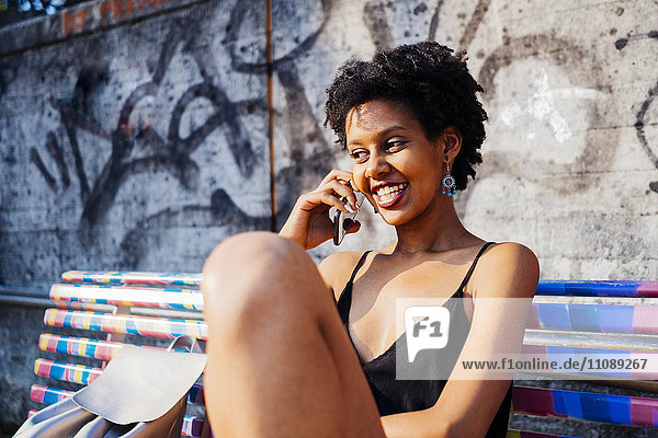 Porträt einer glücklichen jungen Frau  die auf einer Bank sitzt und mit dem Handy telefoniert.