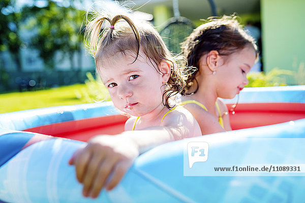 Portrait of baby girl sitting in paddling pool with her sister in the background