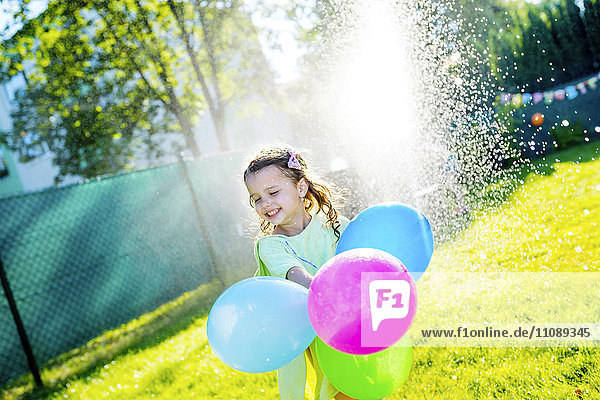 Little girl with balloons having fun with lawn sprinkler in the garden