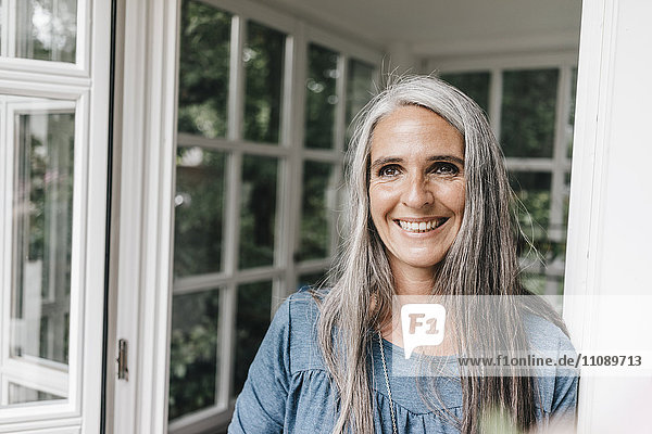 Porträt einer glücklichen Frau mit langen grauen Haaren  die sich an den Türrahmen ihres Wintergartens lehnt.
