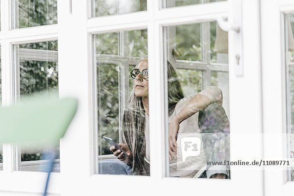 Nachdenkliche Frau mit Telefongespräch im Wintergarten mit Blick durchs Fenster