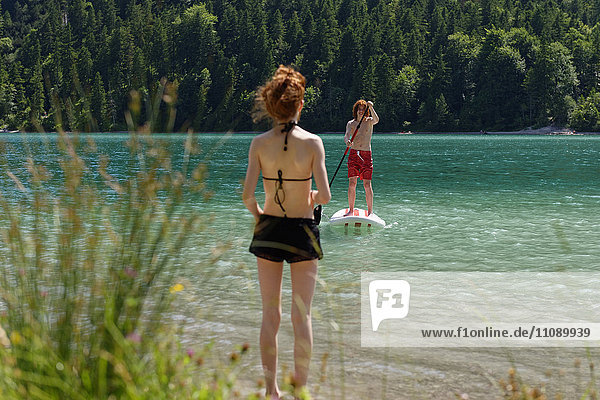 Österreich  Tiroler Ammergauer Alpen  Plansee  Teenagerjunge auf dem Paddelbrett stehend  Schwester am Seeufer wartend