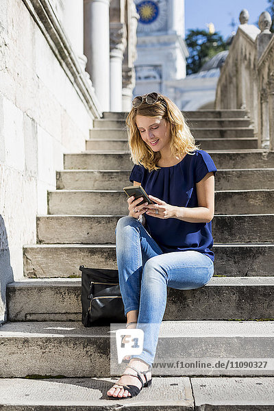 Italien  Udine  lächelnde blonde Frau sitzt auf der Treppe und schaut aufs Handy