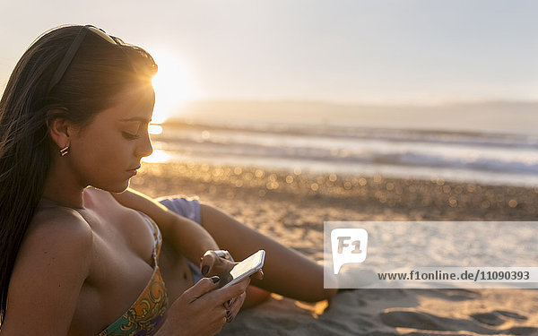 Spanien  Asturien  schöne junge Frau mit Smartphone am Strand bei Sonnenuntergang