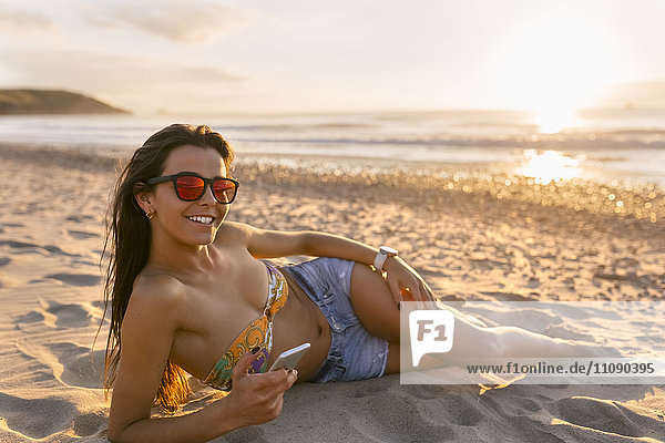 Spain  Asturias  beautiful young woman with smartphone on the beach at sunset