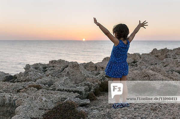 Rückansicht des kleinen Mädchens mit erhobenen Armen an der Felsenküste bei Sonnenuntergang