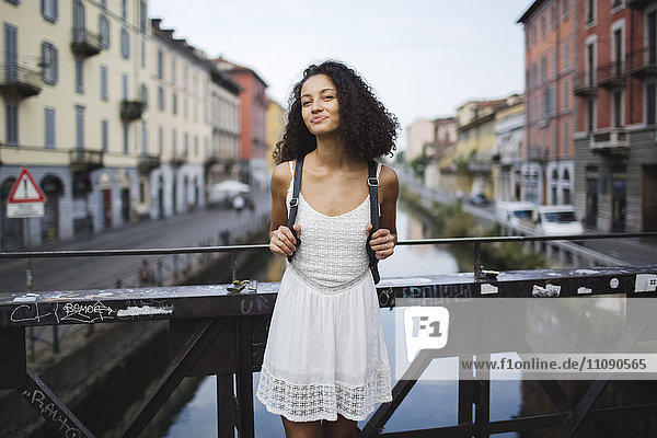 Italien  Mailand  Porträt einer lächelnden jungen Frau mit Rucksack in weißem Sommerkleid auf einer Brücke stehend