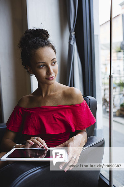 Porträt einer nachdenklichen jungen Frau mit Tablette  die in einem Café sitzt und durchs Fenster schaut.