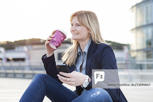 Lächelnde blonde Geschäftsfrau mit Smartphone und Kaffee zum Mitnehmen