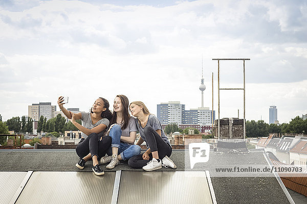 Germany  Berlin  three friends sitting side by side on roof top taking selfie with smartphone