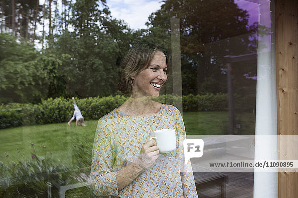 Lächelnde Frau hält Tasse mit Blick aus dem Fenster