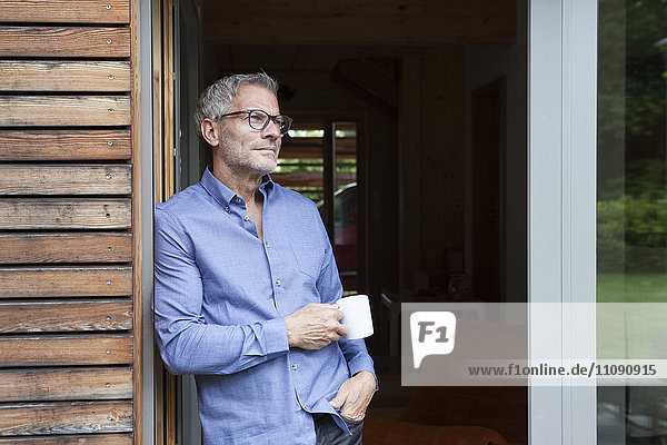 Mature man holding cup leaning against terrace door