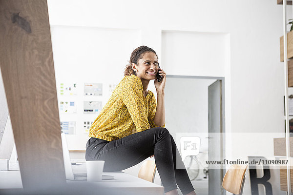 Smiling woman sitting on office desk using cell phone
