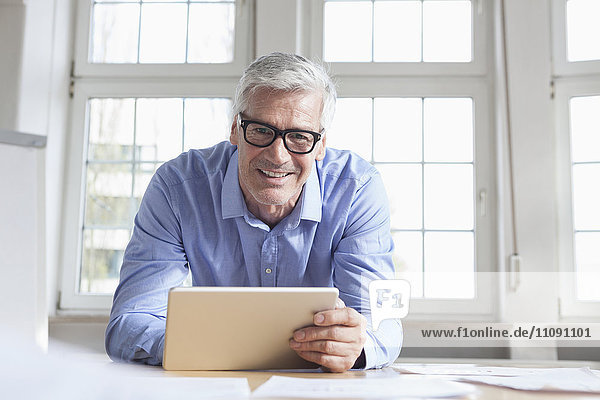 Portrait of smiling mature businessman at office desk holding tablet