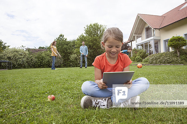 Smiling girl sitting in garden using tablet with family in background