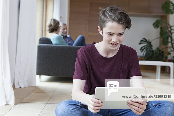 Lächelnder Teenager mit Tablette im Wohnzimmer mit Eltern im Hintergrund