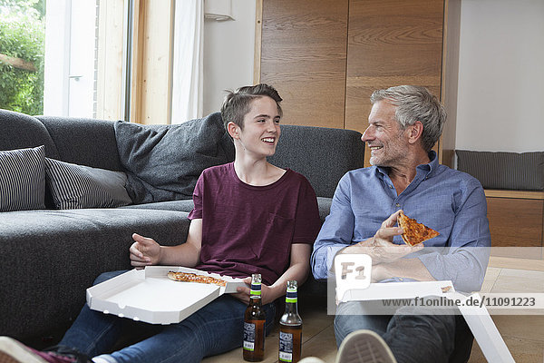 Vater und Sohn sitzen auf dem Boden und essen Pizza im Wohnzimmer.