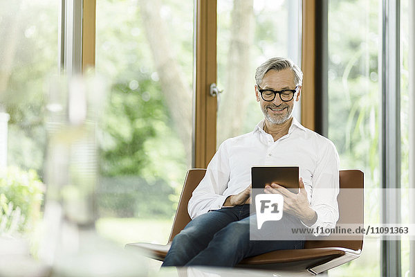 Lächelnder Mann sitzt auf Stuhl in seinem Wohnzimmer mit Tablette