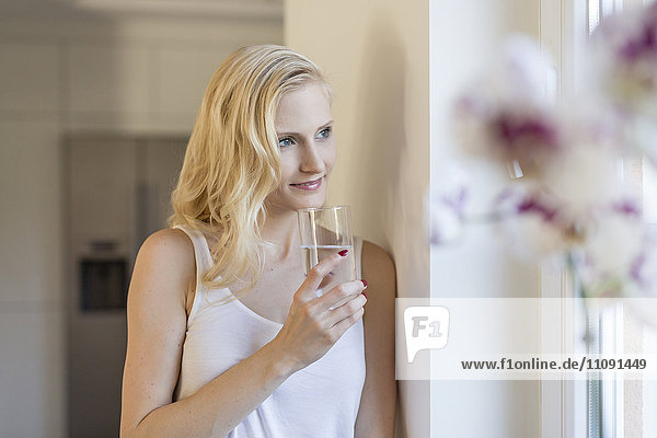 Blonde Frau hält ein Glas Wasser am Fenster.