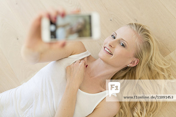 Blond woman lying on wooden floor taking a selfie