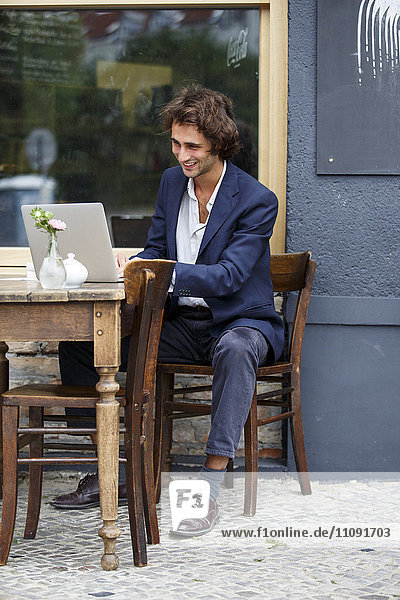 Lächelnder junger Geschäftsmann bei der Arbeit mit dem Laptop im Straßencafé