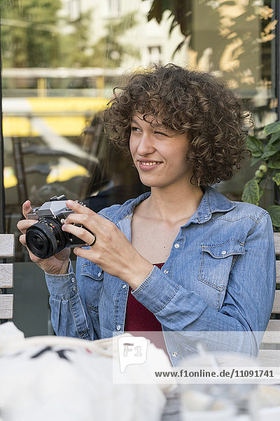 Lächelnde junge Frau mit alter Kamera sitzend in einem Straßencafé