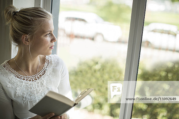 Junge Frau liest ein Buch am Fenster und schaut hinaus.