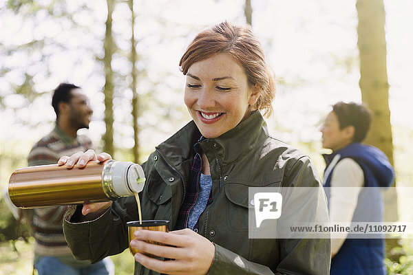 Lächelnde Frau schüttet Kaffee aus einem isolierten Getränkebehälter im Wald aus