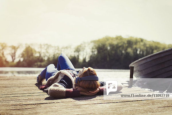 Woman relaxing laying on dock listening to music with headphones at sunny lakeside
