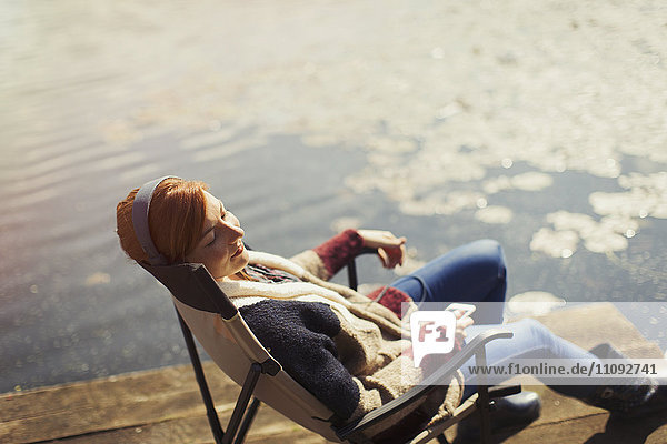 Gelassene Frau entspannt Musik hören mit Kopfhörer und Smartphone am sonnigen Seeufer Dock