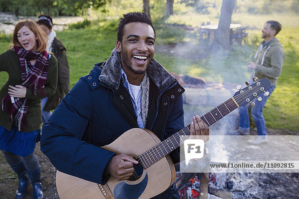 Porträt eines lächelnden Mannes  der mit Freunden auf dem Campingplatz Gitarre spielt.