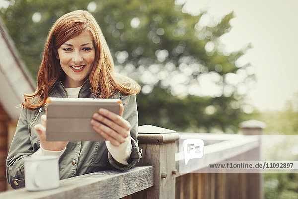 Smiling woman with red hair using digital tablet and drinking coffee at balcony railing