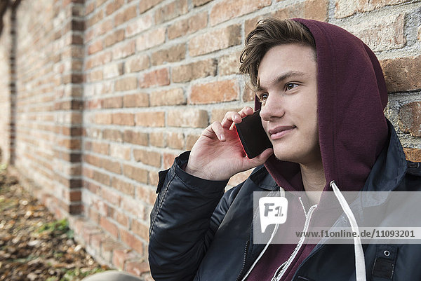 Junger Mann spricht mit einem Handy und lehnt sich an eine Backsteinmauer