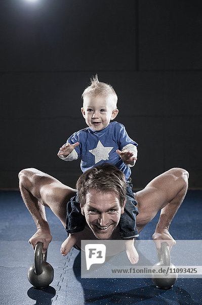 Porträt eines lächelnden Sportlers  der Liegestütze macht und seinen Sohn auf der Schulter trägt