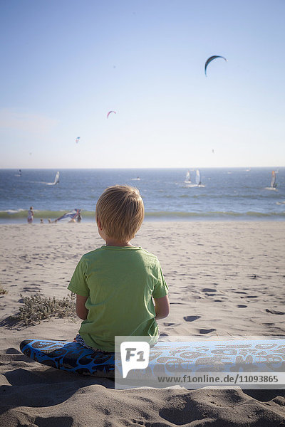 Rückansicht eines kleinen Kindes  das die Aussicht betrachtet und am Strand sitzt