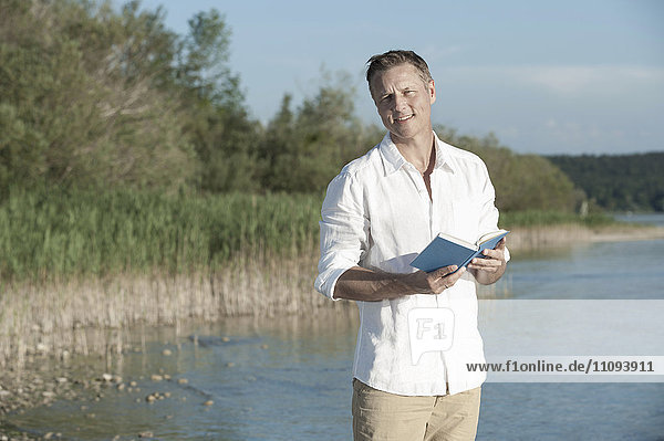 Mature man reading book and smiling at lakeside  Bavaria  Germany