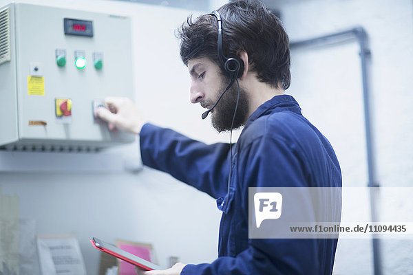 Junger männlicher Ingenieur mit Headset  der mit Hilfe eines digitalen Tablets in einer Industrieanlage die Steuertafel aktualisiert