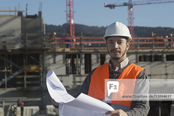 Porträt eines jungen männlichen Ingenieurs  der auf einer Baustelle einen Bauplan hält