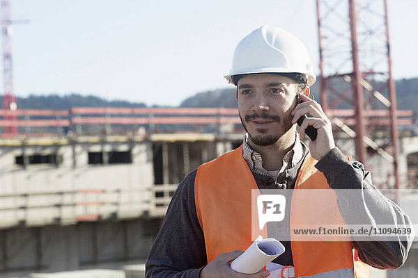 Junger männlicher Ingenieur  der einen Bauplan hält und auf einer Baustelle mit einem Mobiltelefon spricht
