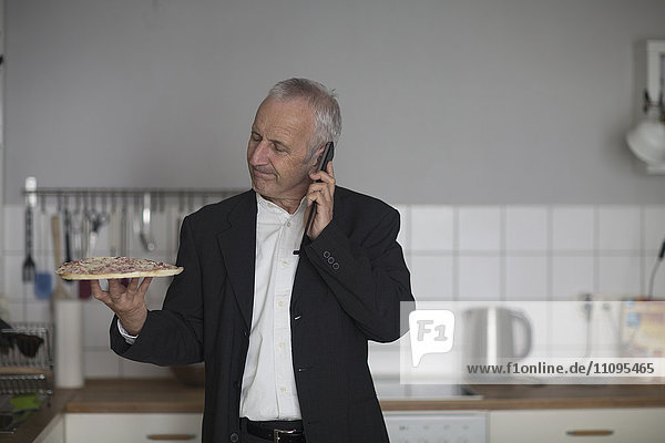 Senior businessman eating pizza and talking on mobile phone in the kitchen  Freiburg im Breisgau  Baden-Württemberg  Germany