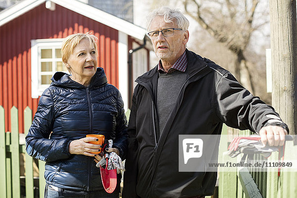 Smiling senior couple holding gardening equipment while standing against fence during winter