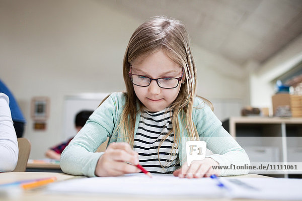 Konzentriertes Mädchen mit Brille beim Schreiben am Klassenzimmer-Schreibtisch in der Schule