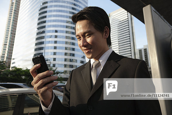 Asiatischer Geschäftsmann  der im Freien eine Textnachricht auf seinem Mobiltelefon sendet
