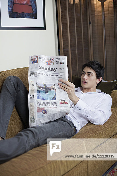 Gemischtrassiger Mann liegt auf dem Sofa und liest Zeitung